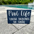 A rectangular decorative plaque with the text 'Pool Life ‘cause beaches be salty' placed on a poolside pavement.