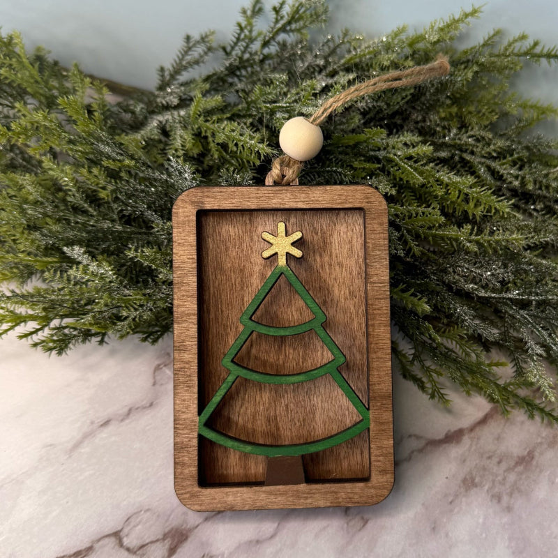 Wooden Christmas tree ornament with green tree design and gold star on a marble surface with greenery in the background.