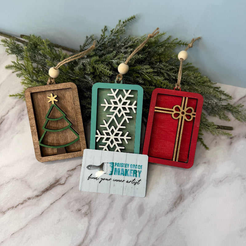 Three wooden Christmas ornaments with a tree, snowflake, and gift box design on a marble surface with greenery.
