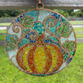 A circular stained glass hanging ornament featuring a ribbed pumpkin in yellow, orange, and red tones, surrounded by green leaves and swirling vines on a blue and teal background, suspended by a loop from a weathered wooden fence over a grassy outdoor setting.
