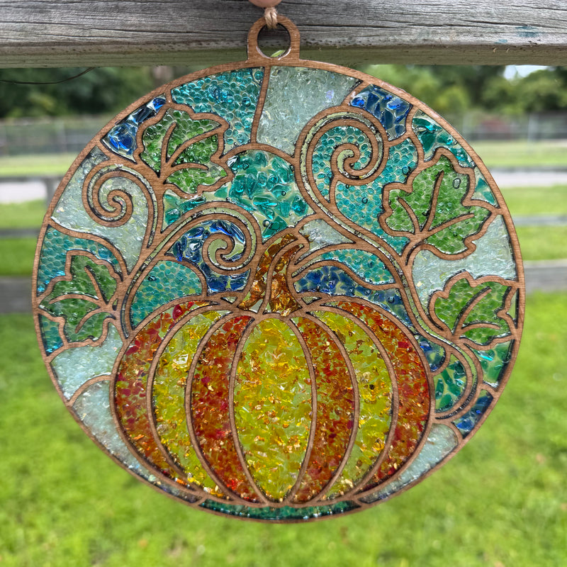 A circular stained glass hanging ornament featuring a ribbed pumpkin in yellow, orange, and red tones, surrounded by green leaves and swirling vines on a blue and teal background, suspended by a loop from a weathered wooden fence over a grassy outdoor setting.