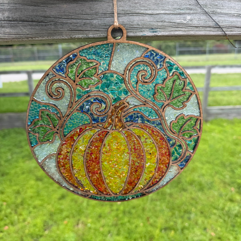 A circular stained-glass-style hanging ornament featuring a pumpkin in yellow, orange, and green, with swirling vine motifs and a rustic copper-toned border, suspended outdoors against a blurred grassy background.