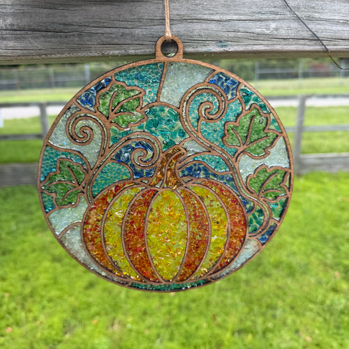 A circular stained-glass-style hanging ornament featuring a pumpkin in yellow, orange, and green, with swirling vine motifs and a rustic copper-toned border, suspended outdoors against a blurred grassy background.
