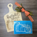 Wooden cutting board with 'Pumpkin Season' engraving, blue card with pumpkin design, and orange ribbon on a wooden surface.