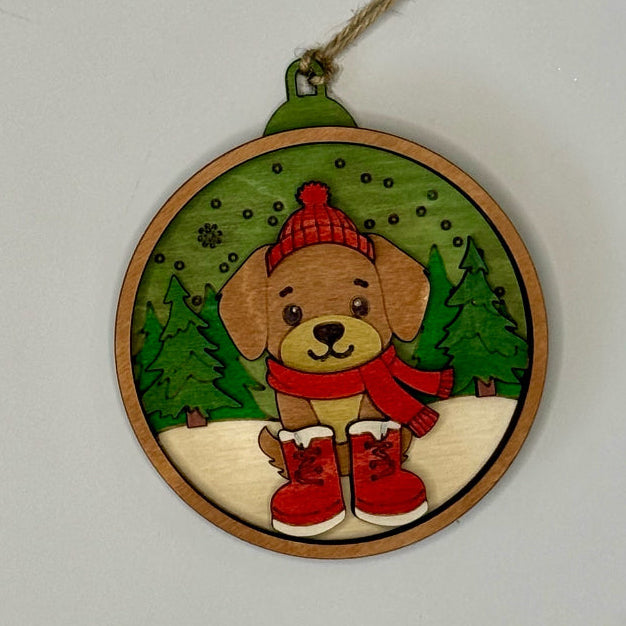 Decorative Christmas ornament with a dog wearing a red hat and scarf, standing on snow with trees in the background.