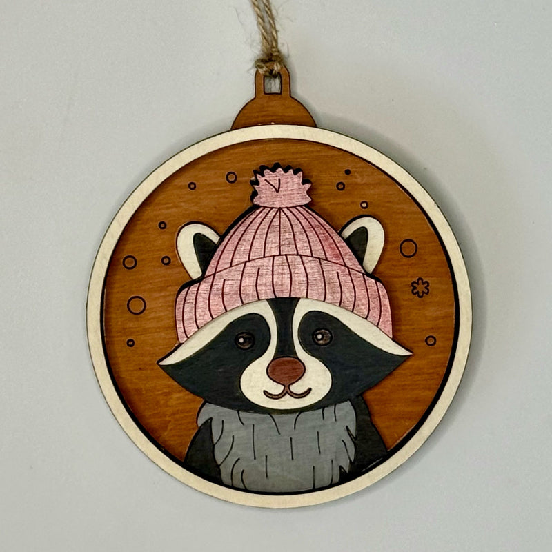 Decorative wooden ornament with a raccoon wearing a pink beanie on a plain background