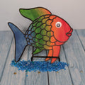 A colorful metal sculpture of a Rainbow Fish standing on a platform with a blue and multicolored body, red fins, and a green and blue scaly texture.