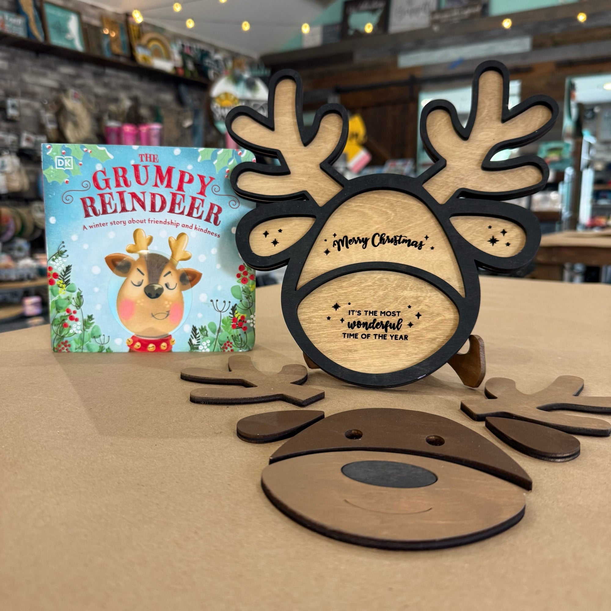 A wooden Christmas reindeer face ornament with antlers and a small children's book titled 'The Grumpy Reindeer' displayed on a tabletop in a shop setting.
