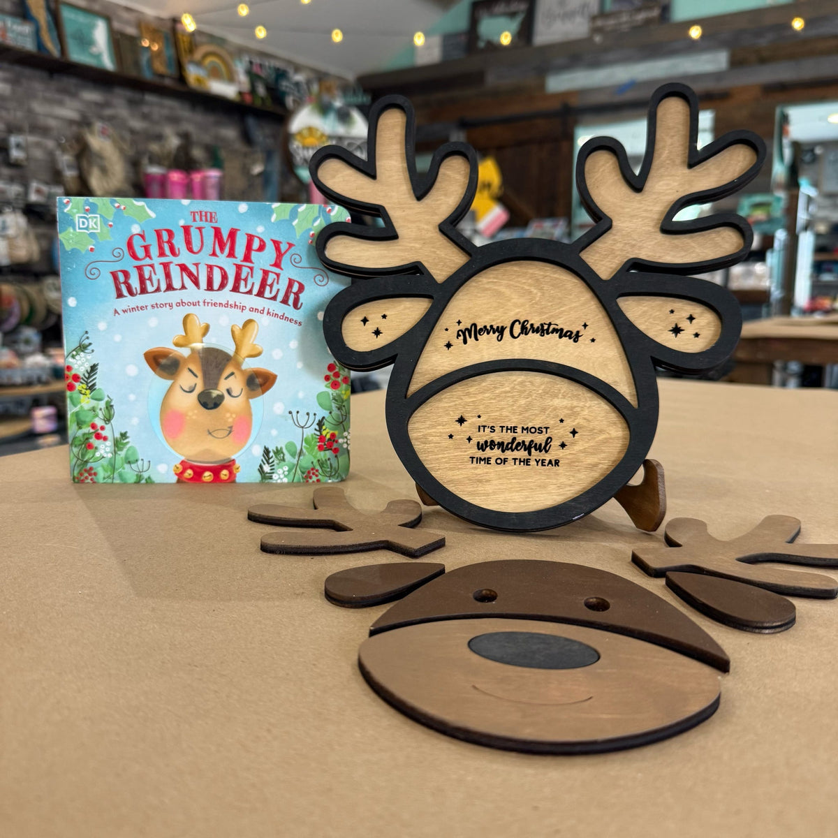 A wooden Christmas reindeer face ornament with antlers and a small children's book titled 'The Grumpy Reindeer' displayed on a tabletop in a shop setting.