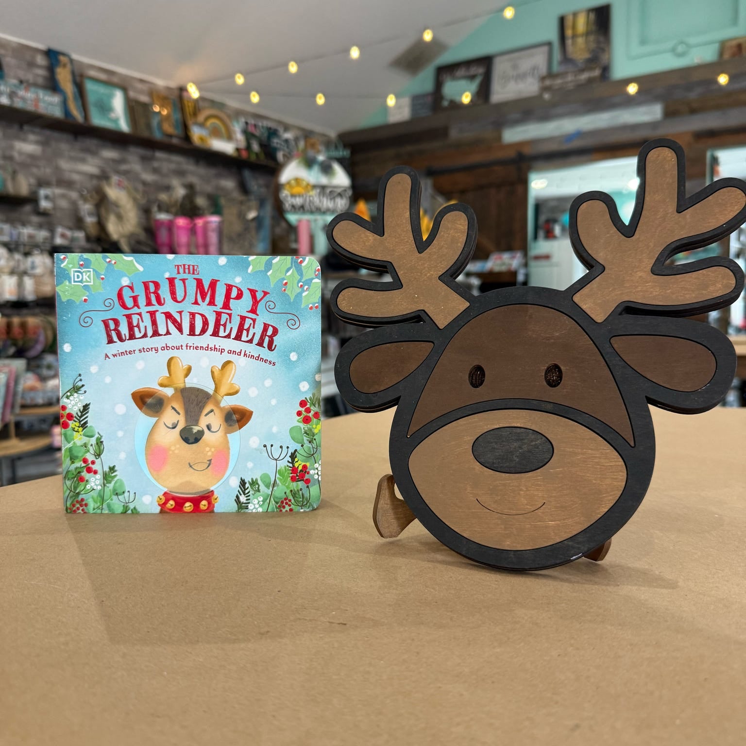 A wooden Christmas-themed standee shaped like a reindeer head with antlers stands next to a children's book titled 'The Grumpy Reindeer' on a tabletop in a shop setting.