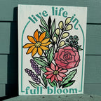 Decorative sign with floral design and 'live life in full bloom' text on a wooden surface.