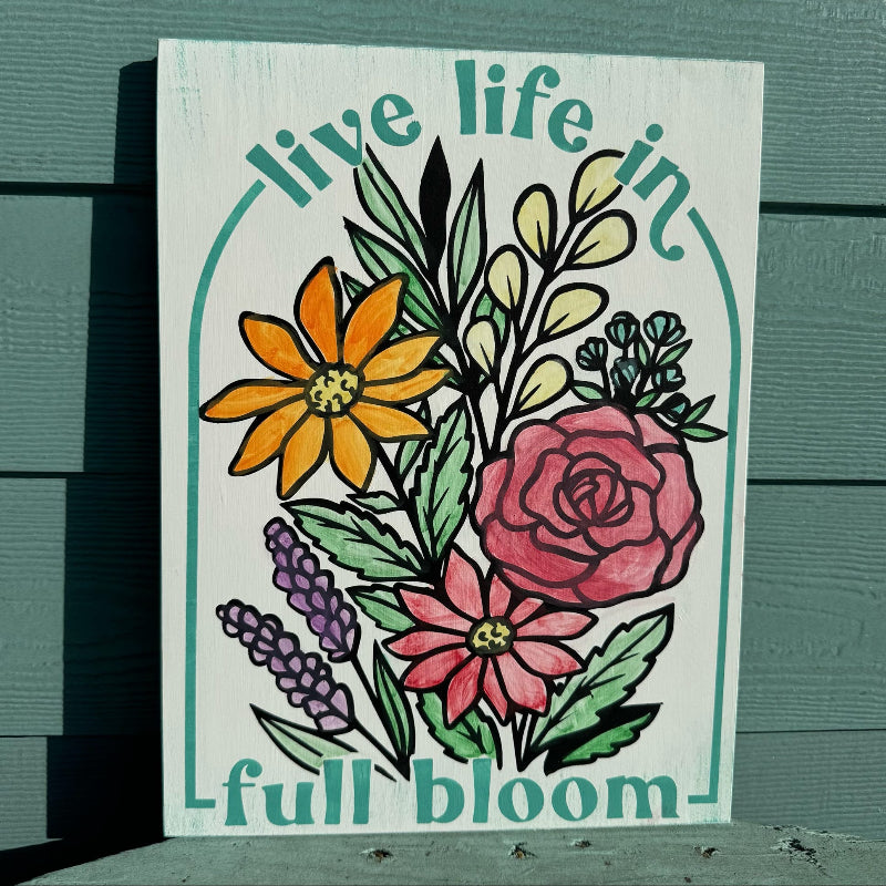 Decorative sign with floral design and 'live life in full bloom' text on a wooden surface.