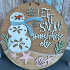 Wooden circular sign with a snowman design and 'Let it snow somewhere else' text, against a wooden background.