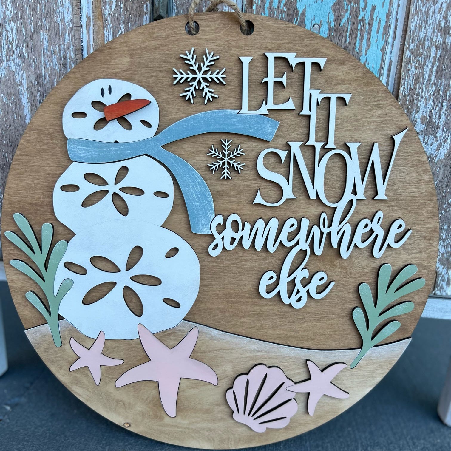 Wooden circular sign with a snowman design and 'Let it snow somewhere else' text, against a wooden background.