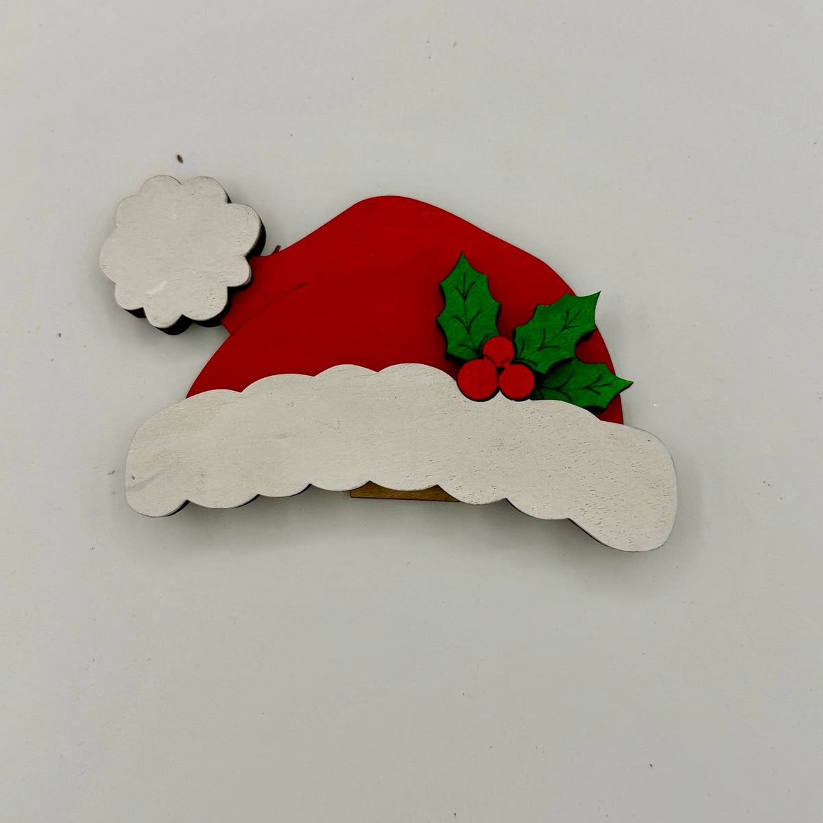 Decorative Christmas ornament with a red Santa hat, white beard, and holly leaves on a light gray background.