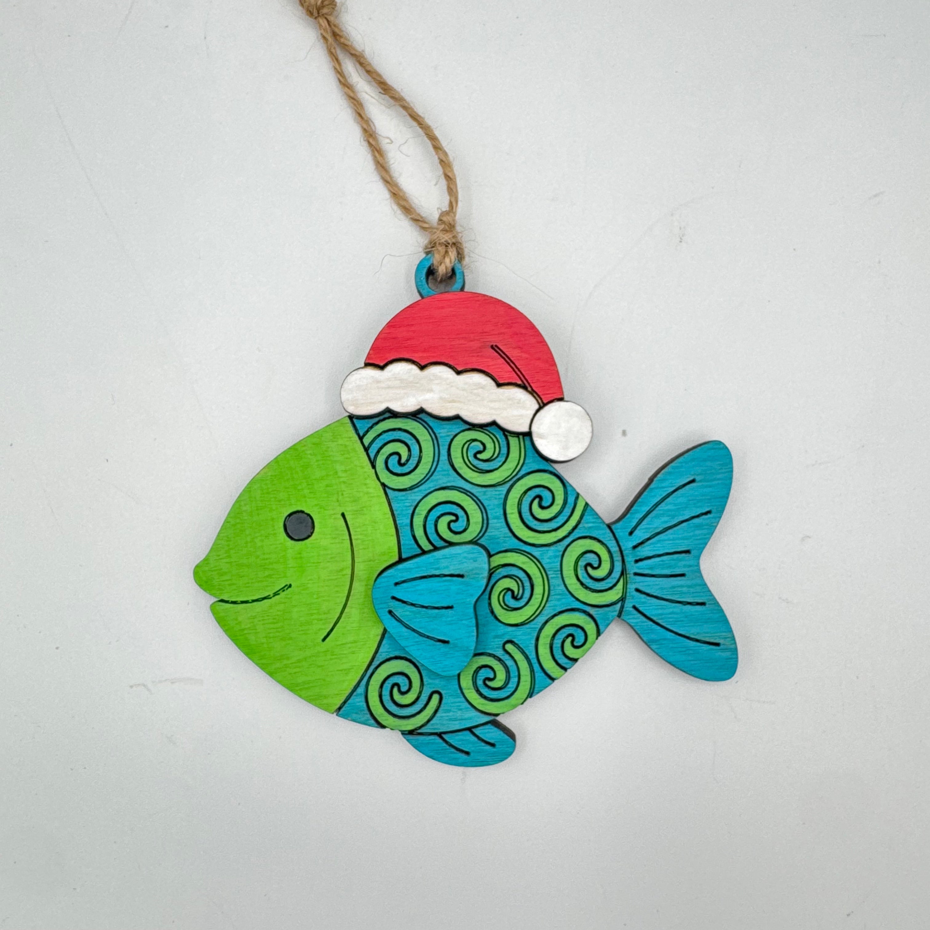 A hand-painted wooden Christmas fish ornament with a green head, blue and green spiral patterned body, a red Santa hat, and a jute hanging string, shown on a plain light gray background.