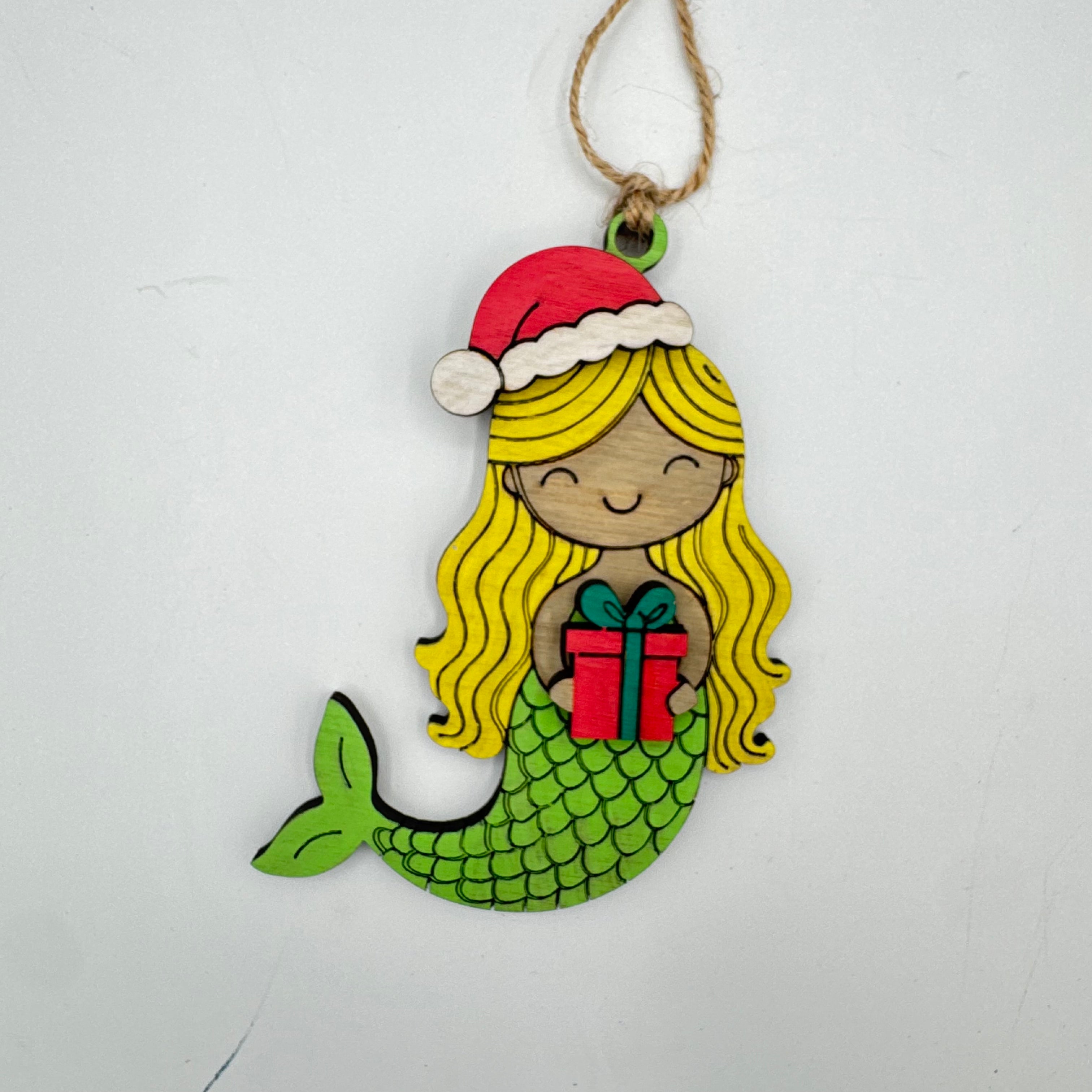 A handcrafted wooden Christmas ornament featuring a mermaid with long yellow hair, a green scaly tail, a red Santa hat, and a red gift box, hanging from a jute twine string against a plain white background.