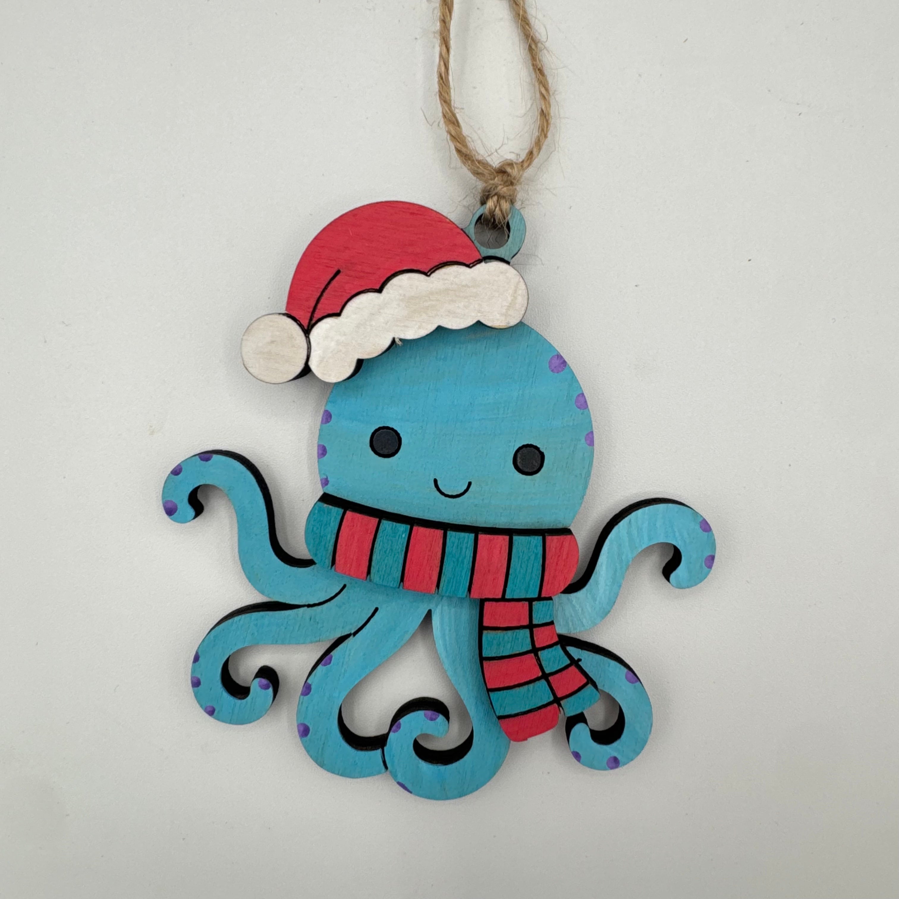 A handcrafted wooden octopus Christmas ornament painted in light blue, wearing a red Santa hat with white trim and a red and blue striped scarf, hanging from a natural jute twine.