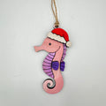 A handcrafted wooden seahorse ornament painted pink and purple, wearing a red and white Santa hat, hanging from a jute twine string against a light gray background.