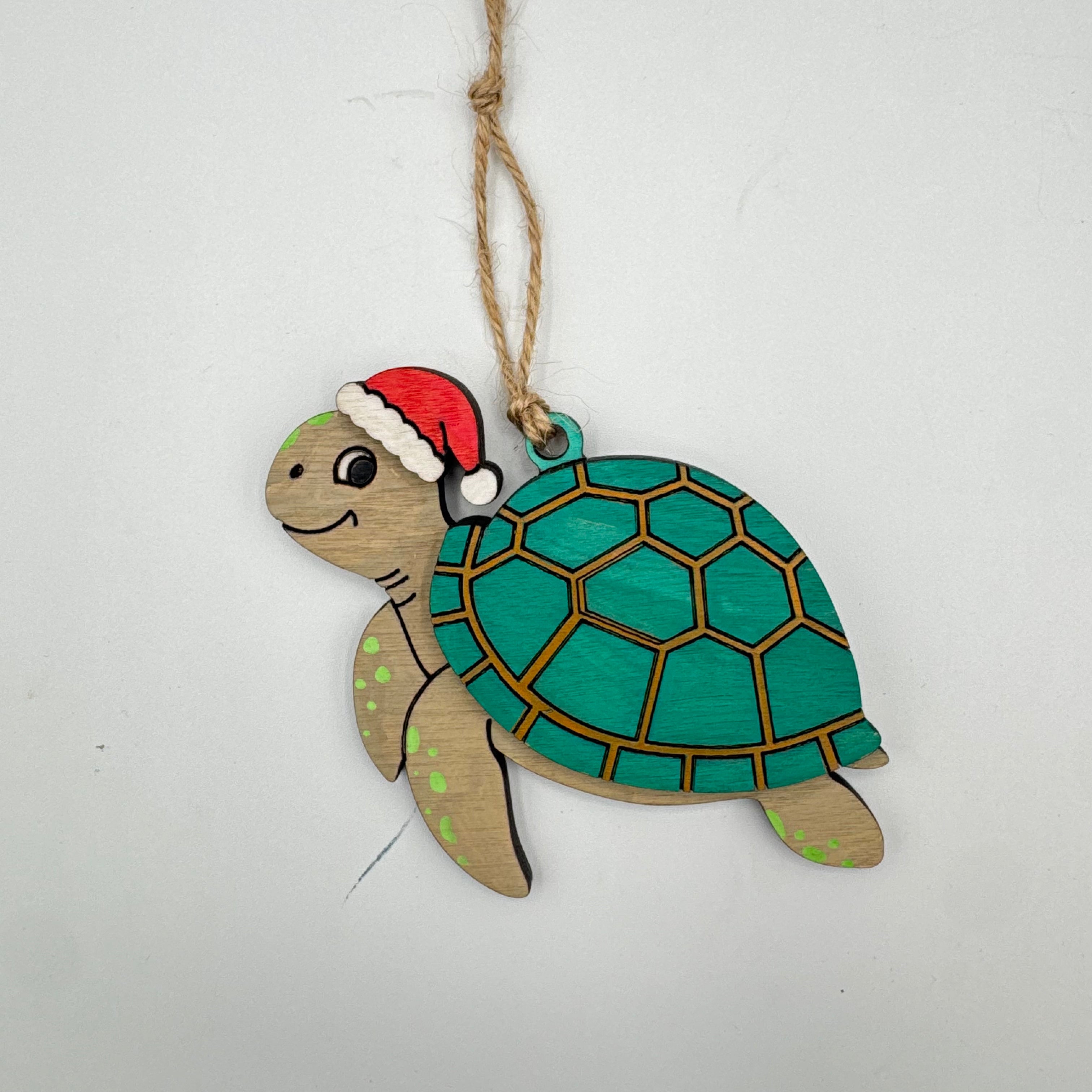 A wooden turtle Christmas ornament painted with a teal green shell, light brown legs, and a red Santa hat, hanging from a natural jute twine string against a plain white background.