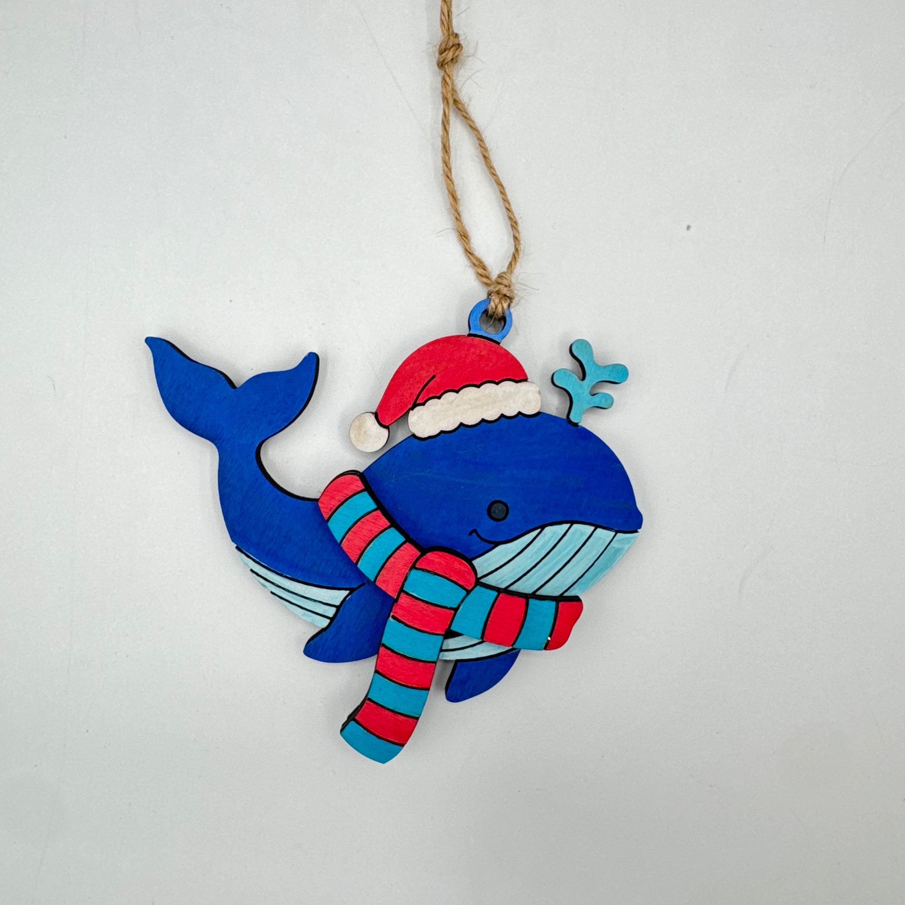 A handcrafted wooden whale Christmas ornament painted in blue, red, and light blue, wearing a red Santa hat and striped red and blue scarf, hanging from a jute twine string against a plain white background.