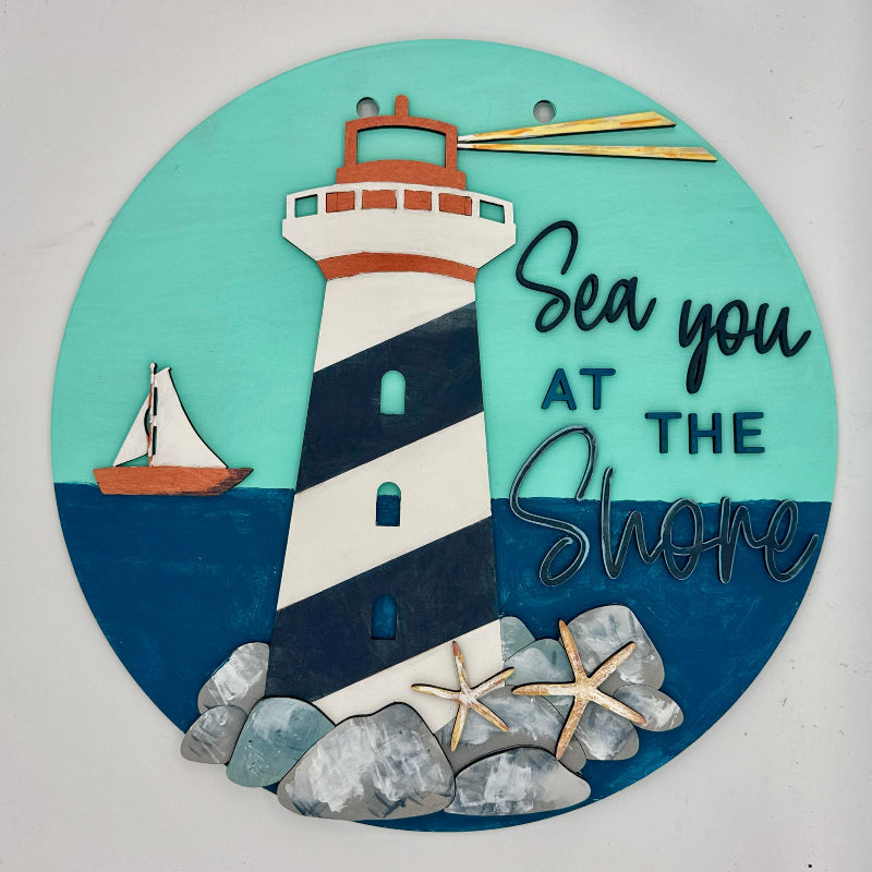 Decorative sign with a lighthouse, sailboat, and 'Sea you at the Shore' text on a teal background.
