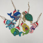 A set of seven colorful wooden Christmas sea-themed ornaments, including a whale, turtle, octopus, mermaid, seahorse, and fish, each wearing a Santa hat and scarf, hanging from twine on a white background.