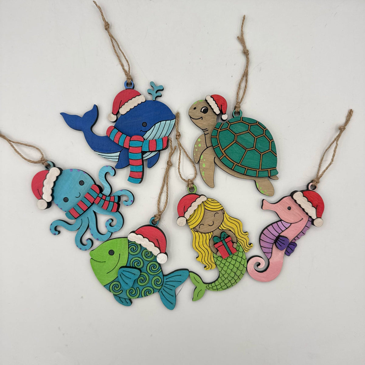 A set of seven colorful wooden Christmas sea-themed ornaments, including a whale, turtle, octopus, mermaid, seahorse, and fish, each wearing a Santa hat and scarf, hanging from twine on a white background.