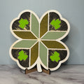 A quilt with a shamrock pattern, featuring green shamrocks on a brown background with gold accents, displayed on a stand.
