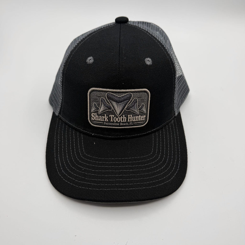 A black and gray trucker hat with a rectangular patch on the front that reads 'Shark Tooth Hunter'. The front panel and brim are black, while the back mesh is gray.