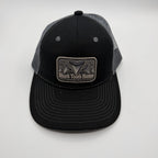 A black and gray trucker hat with a rectangular patch on the front that reads 'Shark Tooth Hunter'. The front panel and brim are black, while the back mesh is gray.
