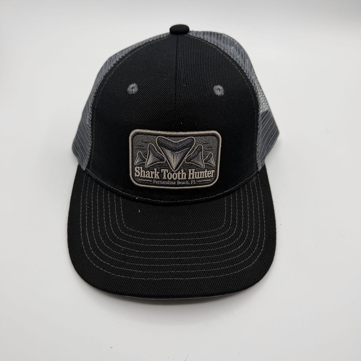 A black and gray trucker hat with a rectangular patch on the front that reads 'Shark Tooth Hunter'. The front panel and brim are black, while the back mesh is gray.