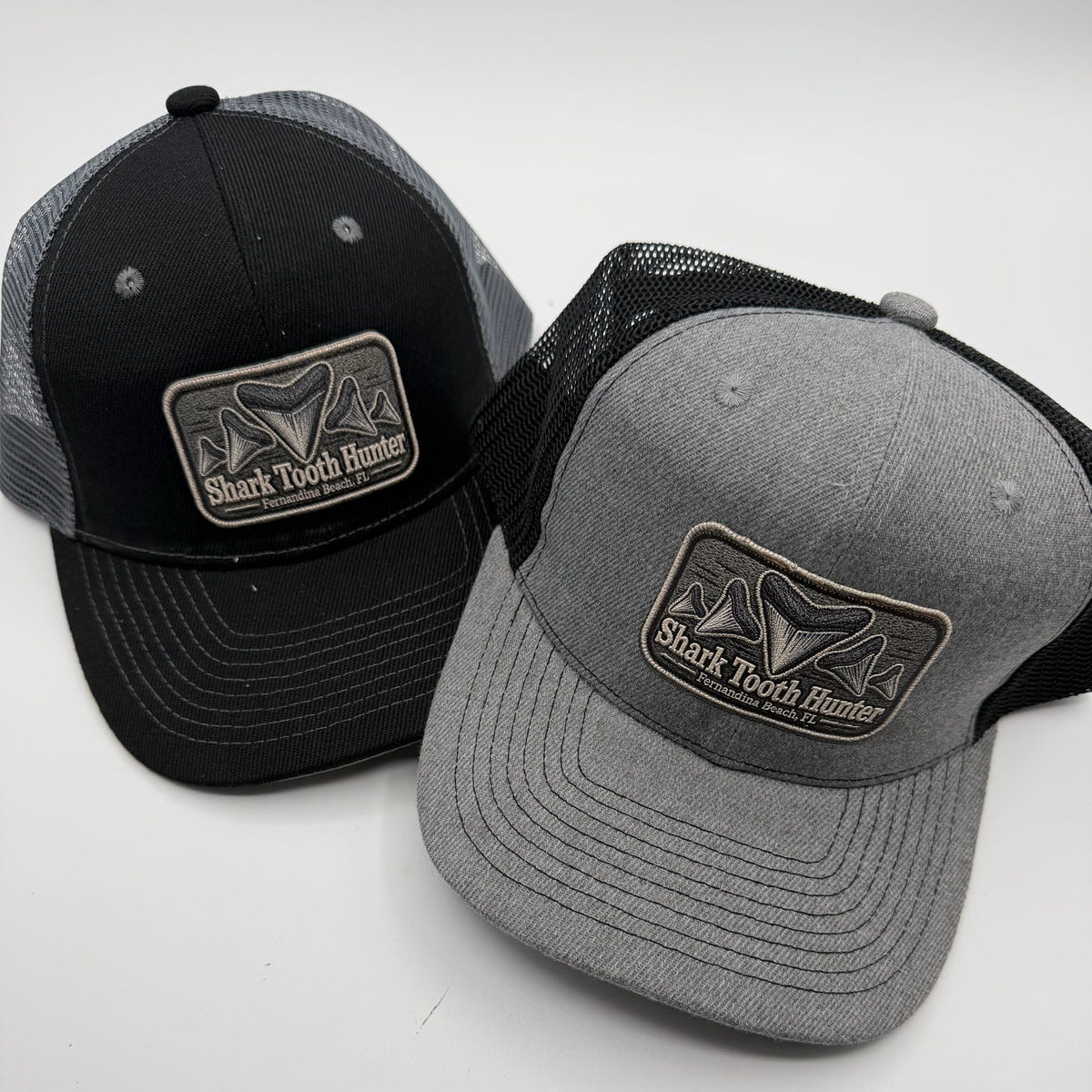 Two trucker hats on a light gray background, one black and one heather gray, both with a rectangular front patch reading Shark Tooth Hunter.