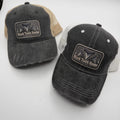 Two distressed trucker hats on a light gray background. The left hat is washed charcoal with beige mesh back panels and a 'Shark Tooth Hunter' patch.