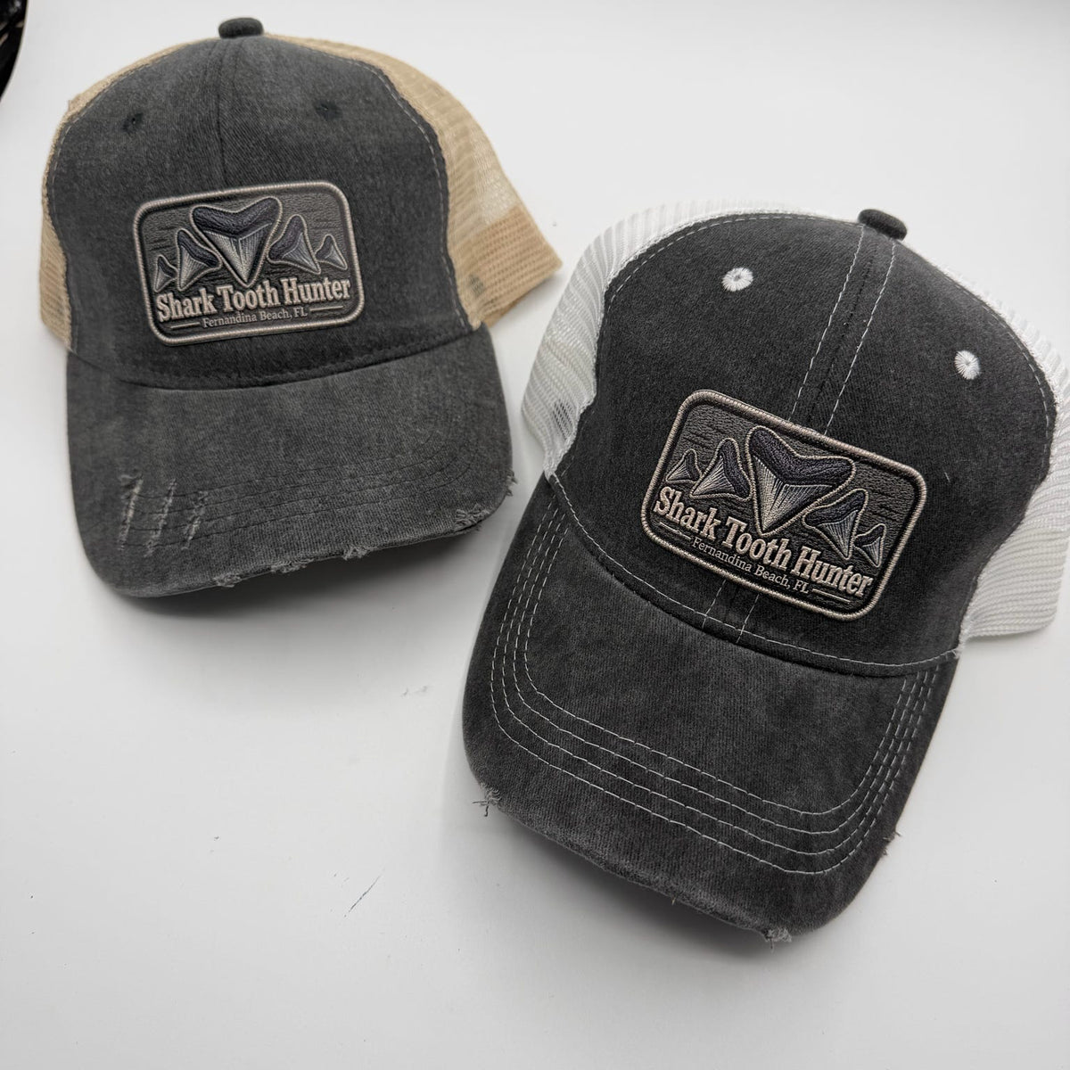 Two distressed trucker hats on a light gray background. The left hat is washed charcoal with beige mesh back panels and a 'Shark Tooth Hunter' patch.