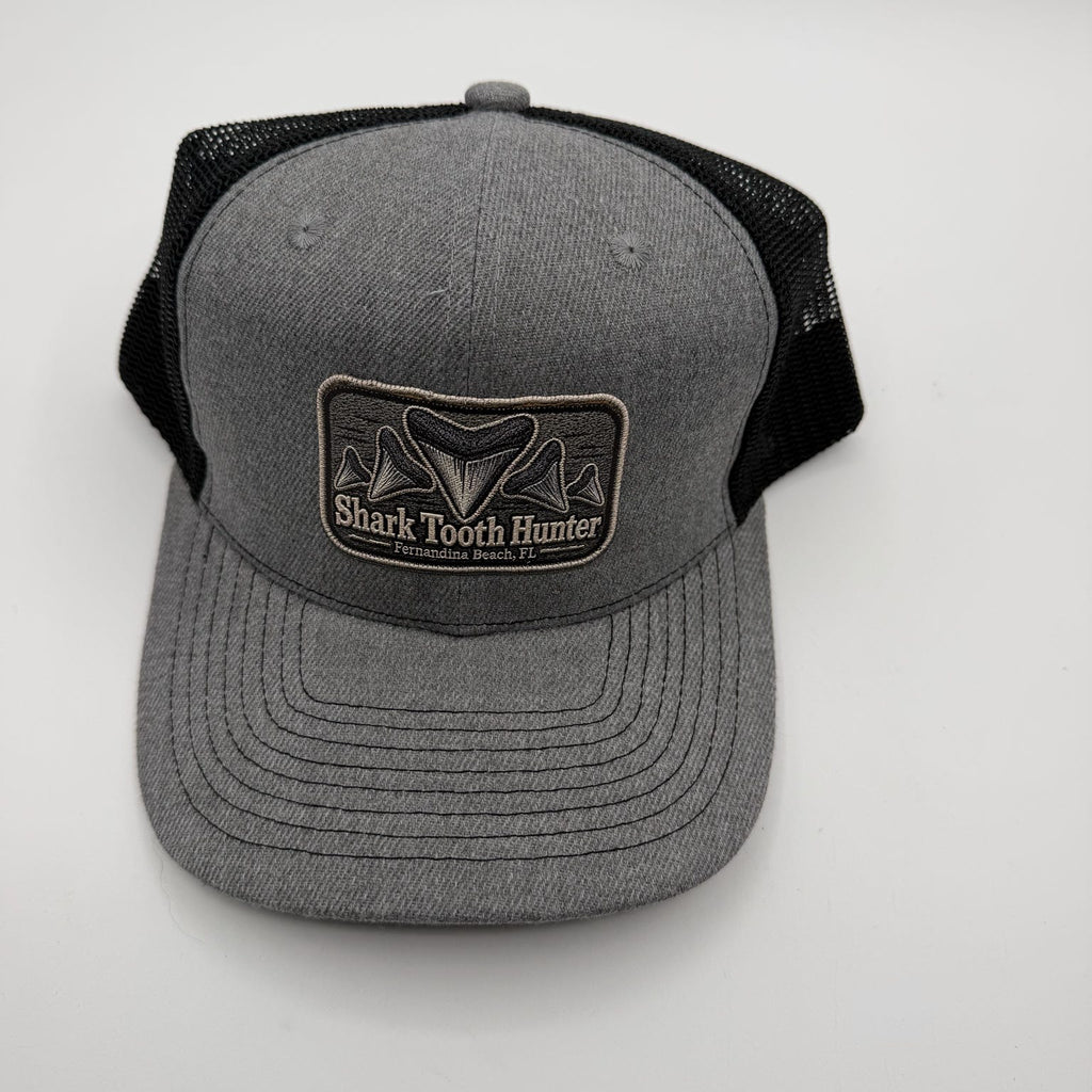 A gray and black trucker hat with a front patch reading 'Shark Tooth Hunter' and a shark fin graphic, photographed on a light gray background.