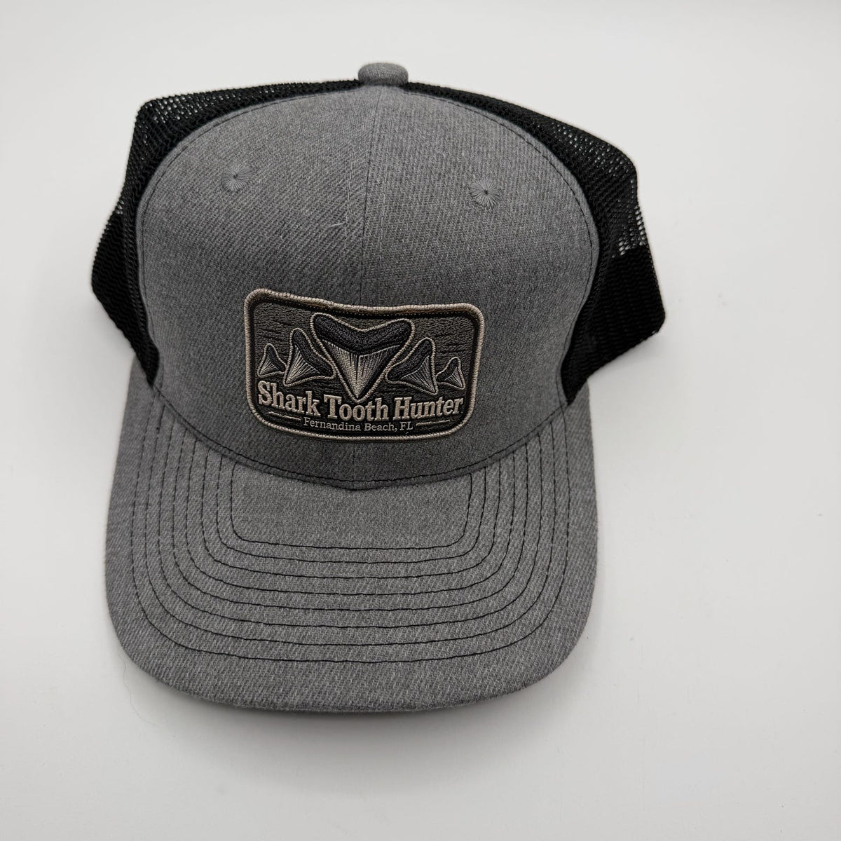A gray and black trucker hat with a front patch reading 'Shark Tooth Hunter' and a shark fin graphic, photographed on a light gray background.