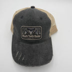 A distressed trucker hat with a dark gray washed denim front panel and brim, and tan mesh back panels. It features a rectangular patch on the front with a shark tooth graphic and the text 'Shark Tooth Hunter'.