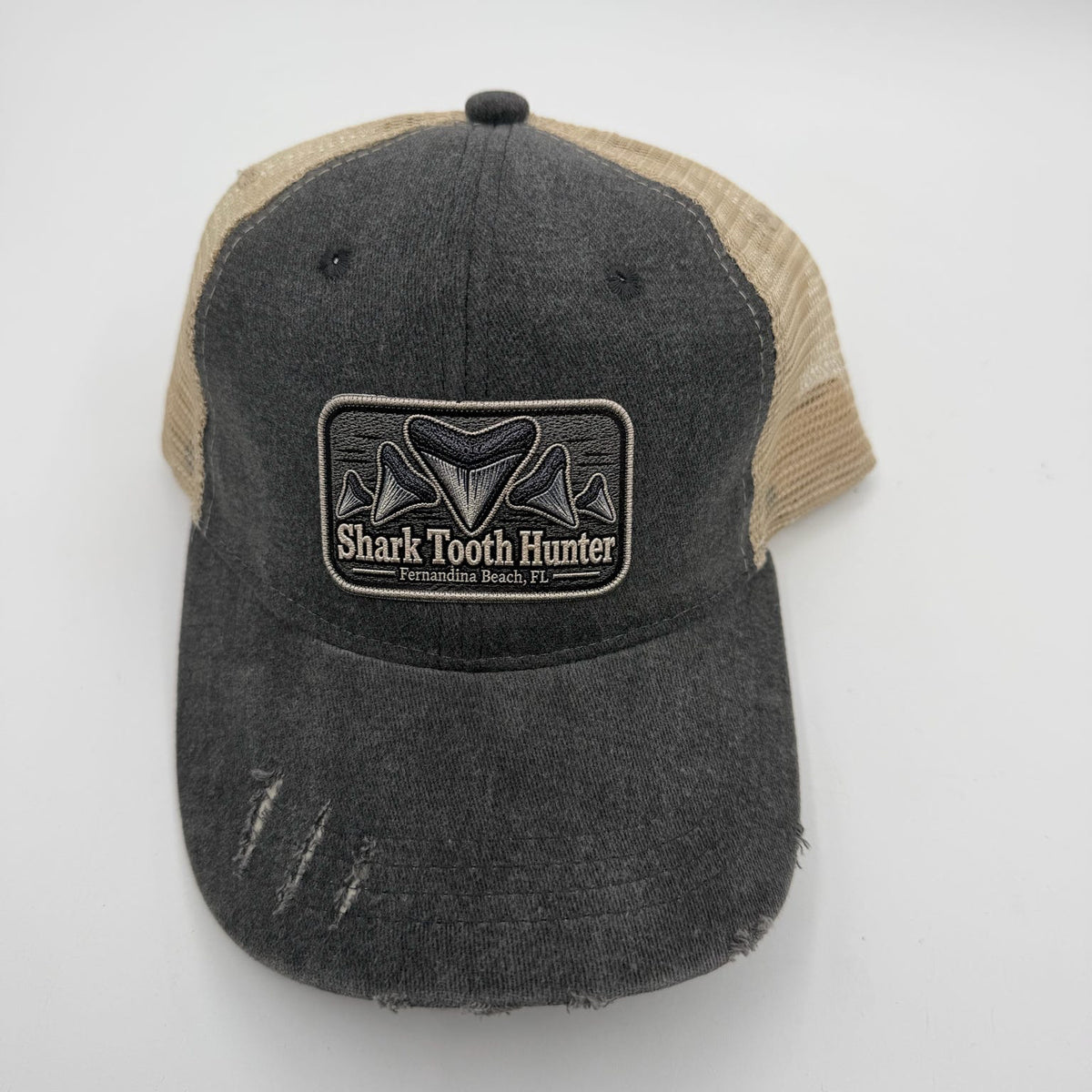 A distressed trucker hat with a dark gray washed denim front panel and brim, and tan mesh back panels. It features a rectangular patch on the front with a shark tooth graphic and the text 'Shark Tooth Hunter'.