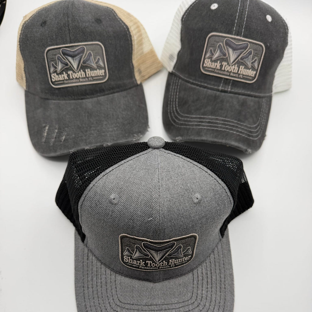 Three trucker-style baseball caps displayed on a white background, each with a rectangular front patch reading Shark Tooth Hunter. The top two caps have dark gray washed fronts with beige and off-white mesh back panels, while the bottom cap has a heather gray front with black mesh back panels.