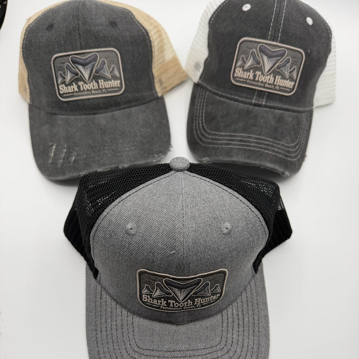 Three trucker-style baseball caps displayed on a white background, each with a rectangular front patch reading Shark Tooth Hunter. The top two caps have dark gray washed fronts with beige and off-white mesh back panels, while the bottom cap has a heather gray front with black mesh back panels.