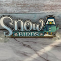 A shelf sitter figurine with the text 'Snow Birds' and a bird design, placed on a marble surface against a wooden backdrop.