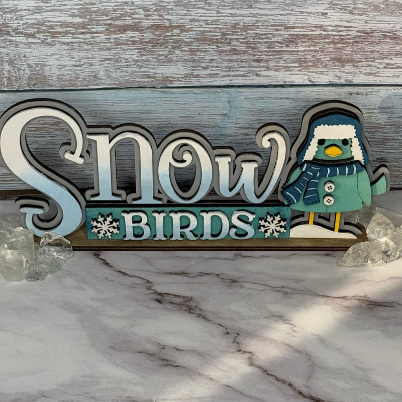 A shelf sitter figurine with the text 'Snow Birds' and a bird design, placed on a marble surface against a wooden backdrop.