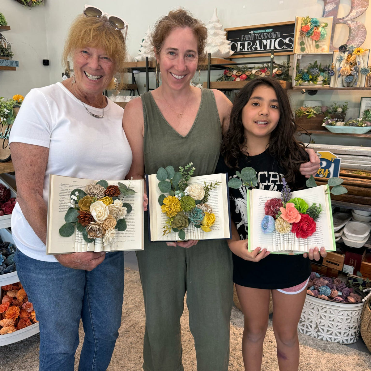 Three women holding floral art pieces in a ceramic studio.