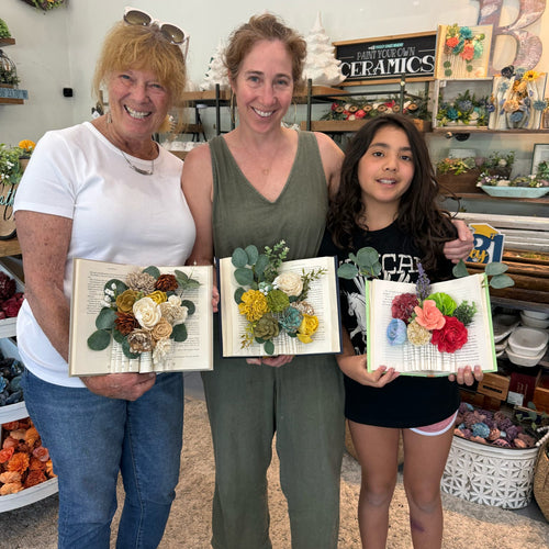 Three women holding floral art pieces in a ceramic studio.