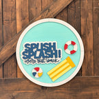 Round decorative sign with 'Splish Splash! We're Out to Lunch' text, beach ball, and life preserver against a wooden background.