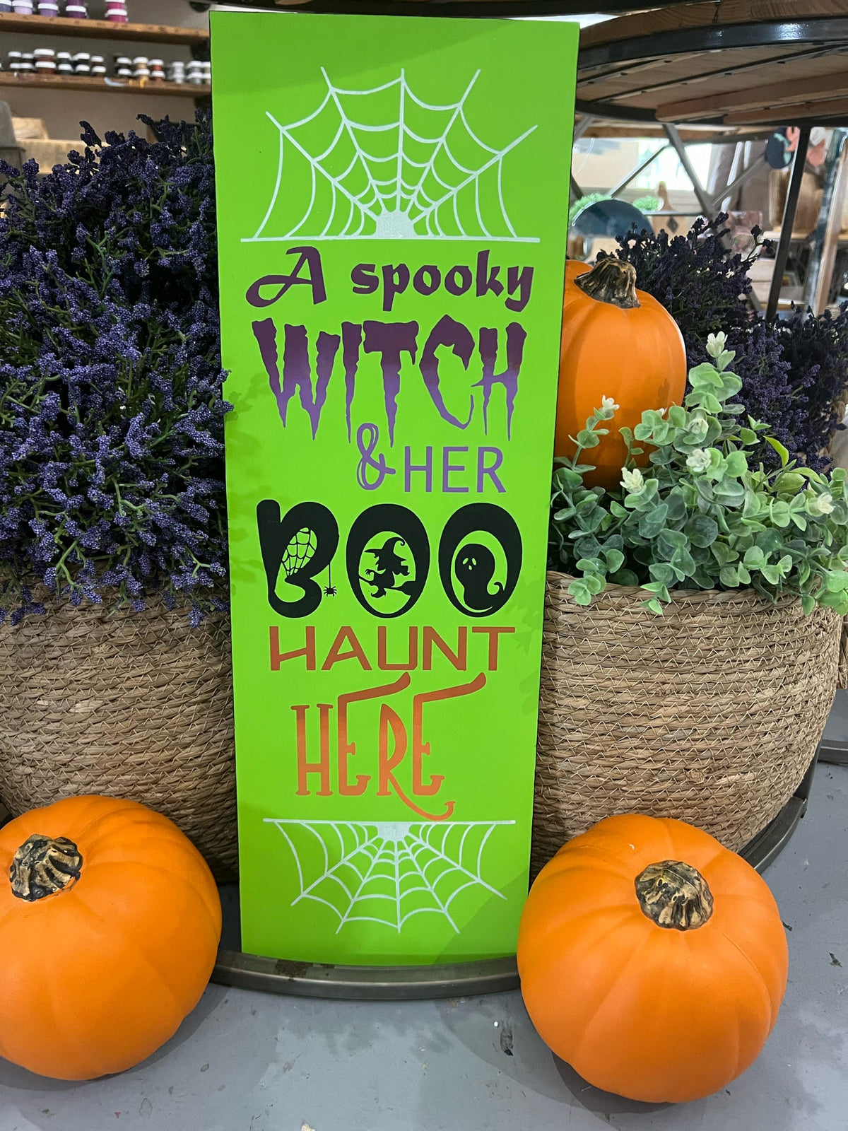 A decorative plank sign featuring a witch and a ghost with the text 'A Spooky Witch & Her Boo HERE' on a green background, displayed in a store with pumpkins and Halloween decorations around it.