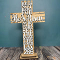 A wooden standing layered cross with the inscription 'He Is Risen' in a cursive script, placed on a wooden surface with a blue background.