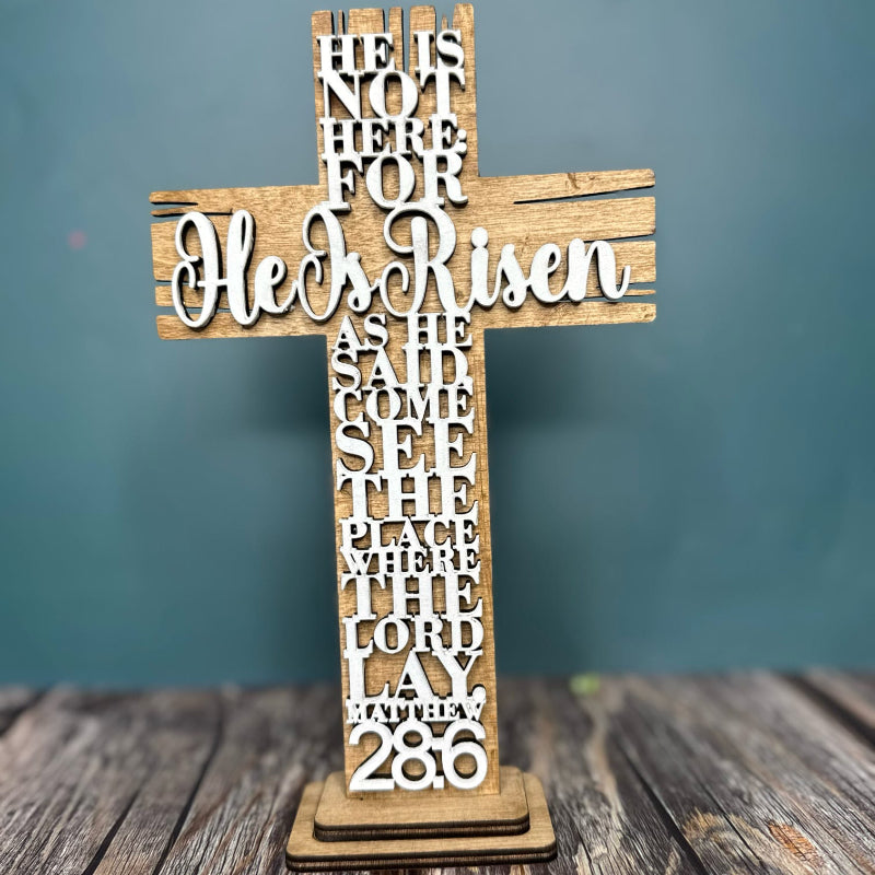 A wooden standing layered cross with the inscription 'He Is Risen' in a cursive script, placed on a wooden surface with a blue background.