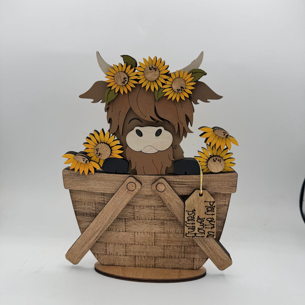 A wooden Highland cow figurine sitting in a basket with sunflowers, on a white background. The cow has brown fur, a white snout, and a yellow flower crown.